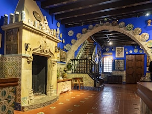 A richly decorated interior features a prominently placed stone fireplace surrounded by ornate ceramic tiles. Shelves are adorned with patterned pottery and vases. The room exhibits a striking deep blue color scheme, complemented by terracotta floor tiles. A staircase leads upward, guarded by wrought iron railings. Decorative plates and artwork enhance the classical ambiance, with a wooden door and arched entryway adding to the historical character.