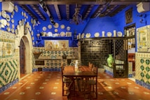 A richly decorated room with vibrant blue walls adorned with numerous decorative plates. The floor is tiled in warm red tones with green and white patterns. A wooden dining table and chairs sit in the center. Intricate tiled designs cover the walls and an old-fashioned fireplace with black metal adds character. The ceiling has visible wooden beams and additional pottery and artwork enhance the aesthetic.