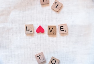 Wooden Scrabble tiles are arranged on a soft, white textile background to spell out the word 'LOVE,' with a small red heart symbol used in place of the letter 'O.' Additional tiles are scattered around, including the letters 'I,' 'T,' 'L,' and 'E.'