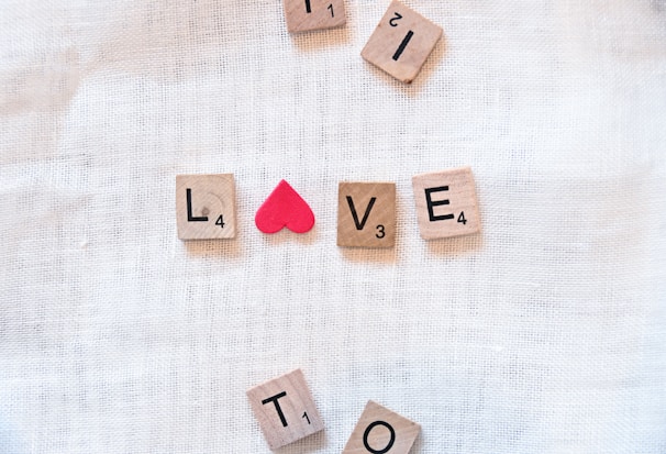 Wooden Scrabble tiles are arranged on a soft, white textile background to spell out the word 'LOVE,' with a small red heart symbol used in place of the letter 'O.' Additional tiles are scattered around, including the letters 'I,' 'T,' 'L,' and 'E.'