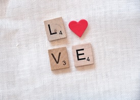 Wooden Scrabble tiles form the word 'LOVE' with a red heart shape positioned above the letter 'O,' arranged on a textured, light-colored fabric background.