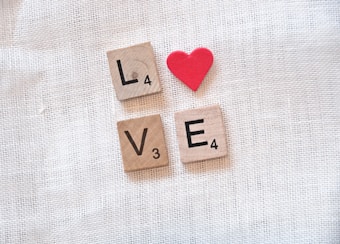 Wooden Scrabble tiles form the word 'LOVE' with a red heart shape positioned above the letter 'O,' arranged on a textured, light-colored fabric background.