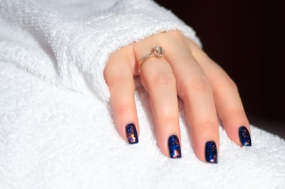 A hand with neatly manicured nails painted in a deep blue shade featuring vibrant sparkles. The hand is adorned with a gold ring set with a gemstone, resting against a fluffy white textured fabric.
