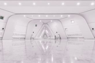A spacious, modern interior with a minimalist design featuring a symmetrical arrangement of curved white walls and a glossy, reflective floor. The space includes small windows, subtle lighting fixtures, and intricate wall details that resemble script or art. At the far end, an artistic installation with geometric patterns is illuminated, adding an artistic focal point.
