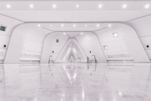 A spacious, modern interior with a minimalist design featuring a symmetrical arrangement of curved white walls and a glossy, reflective floor. The space includes small windows, subtle lighting fixtures, and intricate wall details that resemble script or art. At the far end, an artistic installation with geometric patterns is illuminated, adding an artistic focal point.