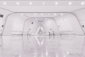 A spacious, modern interior with a minimalist design featuring a symmetrical arrangement of curved white walls and a glossy, reflective floor. The space includes small windows, subtle lighting fixtures, and intricate wall details that resemble script or art. At the far end, an artistic installation with geometric patterns is illuminated, adding an artistic focal point.