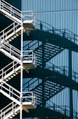A metal staircase with multiple landings and railings casts intricate shadows against a large blue corrugated wall. The geometric patterns create a visually striking interplay between light and shadow.