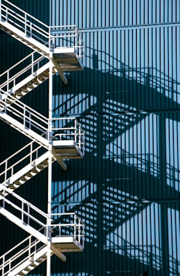 A metal staircase with multiple landings and railings casts intricate shadows against a large blue corrugated wall. The geometric patterns create a visually striking interplay between light and shadow.