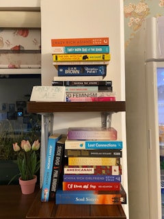 A wooden shelf holding a collection of various books, each with distinct and colorful covers. The books are arranged in two stacks on the shelf. Nearby, a handwritten sign reads 'Only For Sale'. A floral-patterned wall is partially visible in the background, along with a window and a vase of tulips.