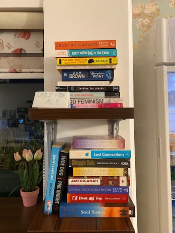A wooden shelf holding a collection of various books, each with distinct and colorful covers. The books are arranged in two stacks on the shelf. Nearby, a handwritten sign reads 'Only For Sale'. A floral-patterned wall is partially visible in the background, along with a window and a vase of tulips.