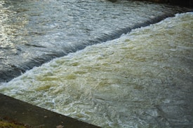 The image captures a section of a river or stream with rushing water, creating a visible contrast between the smooth water surface and the turbulent flow over what appears to be a small dam or weir.