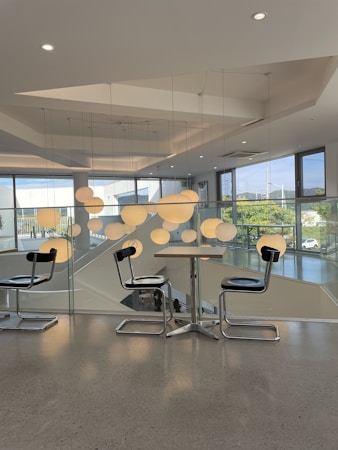 A modern interior with a glass railing and geometric ceiling design. The space features sleek furnishings including a pair of black chairs with chrome legs and a wooden table. Hanging cluster of round pendant lights adds an artistic element. Large windows offer views of the outdoors with greenery and partial blue sky visible.