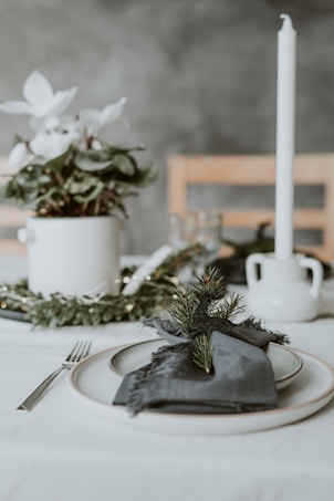 A minimalist table setting features a white plate with a dark cloth napkin, adorned with a small evergreen branch. In the background, there is a white potted plant with white flowers and a tall white candle. The scene is set against a grey wall and includes a wooden chair.