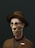 A stylized, digital 3D rendering of a character with a long, angular face wearing round glasses and a brown, striped hat. The character is dressed in a brown textured jacket with a visible name tag reading 'OSCORR'. The background is a solid, dark green, creating a strong contrast with the character's lighter complexion.