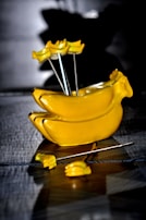 A shiny, yellow banana-shaped object with several banana-shaped picks inserted into it. The surface has a reflective quality and is placed on a textured grey background.