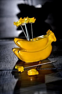 A shiny, yellow banana-shaped object with several banana-shaped picks inserted into it. The surface has a reflective quality and is placed on a textured grey background.