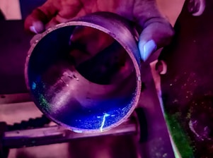 A hand is holding a metallic cylindrical object which is illuminated under a blue and purple light. The lighting highlights the texture and imperfections on the surface of the metal.