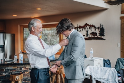An older man assists a younger man with his tie in a warmly lit room. The younger man is wearing a light grey suit, while the older man is in a white shirt and tie. The room features rustic decor, with a wooden table and a wall decoration depicting mountains and a bear silhouette. Bottled water and a cardboard box are visible on a counter in the background.