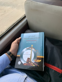 A person is holding a book with an ornate cover featuring a gavel and scales of justice. The book has Arabic text and is resting on a person's lap, who is wearing a light blue shirt and a watch with a black strap. It appears to be in a vehicle, indicated by the seat and window nearby.