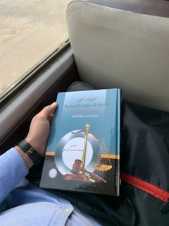 A person is holding a book with an ornate cover featuring a gavel and scales of justice. The book has Arabic text and is resting on a person's lap, who is wearing a light blue shirt and a watch with a black strap. It appears to be in a vehicle, indicated by the seat and window nearby.