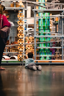 A child lies on the floor gazing at an intricate structure made of metal, wood, and colorful blocks. Nearby, an adult stands with a handbag, observing the structure as well. The scene suggests an environment like a museum or interactive exhibit, with various geometric shapes and textures creating a visually engaging setting.