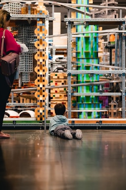 A child lies on the floor gazing at an intricate structure made of metal, wood, and colorful blocks. Nearby, an adult stands with a handbag, observing the structure as well. The scene suggests an environment like a museum or interactive exhibit, with various geometric shapes and textures creating a visually engaging setting.