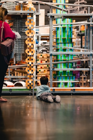 A child lies on the floor gazing at an intricate structure made of metal, wood, and colorful blocks. Nearby, an adult stands with a handbag, observing the structure as well. The scene suggests an environment like a museum or interactive exhibit, with various geometric shapes and textures creating a visually engaging setting.
