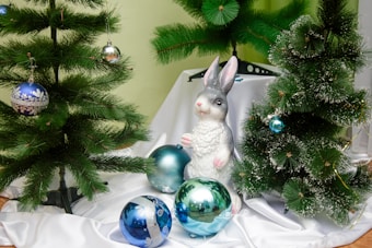 A festive scene with artificial pine trees decorated with blue and silver baubles. In the center, a statue of a rabbit is positioned among the trees, surrounded by additional baubles on a white fabric base. The setting conveys a winter holiday atmosphere, enhanced by the greenery and bright ornaments.