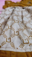A close-up view of a fabric with a geometric pattern consisting of brown square shapes and dot clusters on a beige background. The fabric appears to be part of a garment with visible stitching along the bottom edge. The background is a contrasting mustard color.