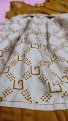A close-up view of a fabric with a geometric pattern consisting of brown square shapes and dot clusters on a beige background. The fabric appears to be part of a garment with visible stitching along the bottom edge. The background is a contrasting mustard color.