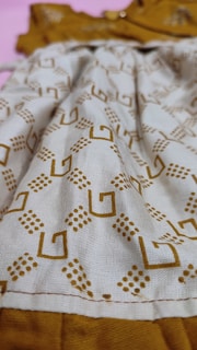 A close-up view of a fabric with a geometric pattern consisting of brown square shapes and dot clusters on a beige background. The fabric appears to be part of a garment with visible stitching along the bottom edge. The background is a contrasting mustard color.