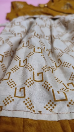A close-up view of a fabric with a geometric pattern consisting of brown square shapes and dot clusters on a beige background. The fabric appears to be part of a garment with visible stitching along the bottom edge. The background is a contrasting mustard color.