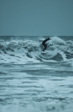 A surfer in a wetsuit rides a wave skillfully against a backdrop of turbulent ocean waters. The waves are large, with frothy white caps, and the sky is overcast, giving the scene a moody and adventurous atmosphere.