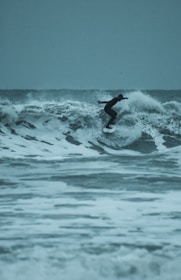 A surfer in a wetsuit rides a wave skillfully against a backdrop of turbulent ocean waters. The waves are large, with frothy white caps, and the sky is overcast, giving the scene a moody and adventurous atmosphere.