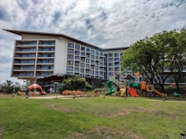 A multi-story modern building with numerous balconies stands adjacent to a well-maintained playground area. The playground features slides, a climbing structure, and colorful play equipment. There are people, including children and adults, engaging in leisure activities. The foreground is a lush green lawn, while the background includes trees and partly cloudy skies.