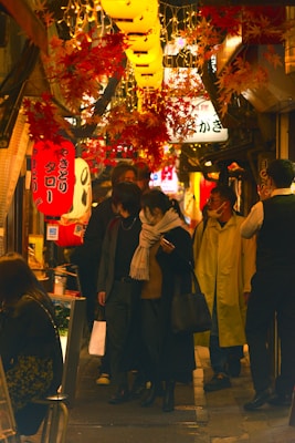 A bustling street scene in a Japanese alley with several people walking and talking. Vibrant red and yellow lanterns, along with autumn leaves, hang above, illuminating the area with a warm glow. Various shop signs are visible in Japanese, contributing to the lively and traditional atmosphere.