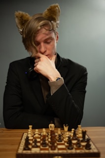 A person wearing a dark blazer and headphones is thoughtfully contemplating a chessboard. They have furry ears on their head, adding a playful touch to the scene. The chessboard in front of them is set up with a mix of black and white chess pieces, suggesting a game in progress.