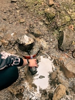 A person filling a plastic bottle from a natural water source in a rocky area. There is moss and greenery growing on the rocks. A vintage-looking water spout is embedded in the rocks.