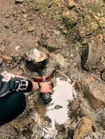 A person filling a plastic bottle from a natural water source in a rocky area. There is moss and greenery growing on the rocks. A vintage-looking water spout is embedded in the rocks.