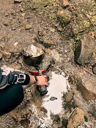 A person filling a plastic bottle from a natural water source in a rocky area. There is moss and greenery growing on the rocks. A vintage-looking water spout is embedded in the rocks.