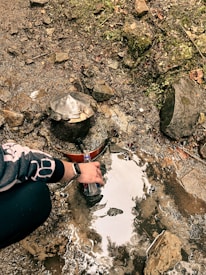 A person filling a plastic bottle from a natural water source in a rocky area. There is moss and greenery growing on the rocks. A vintage-looking water spout is embedded in the rocks.