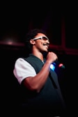 A person with curly hair and a mustache is holding a microphone, wearing sunglasses and a dark sleeveless sweater over a white shirt. The stage is dimly lit, creating a dramatic and focused atmosphere.