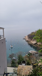 A tranquil coastal scene with a sailboat in a calm, clear water bay. The shoreline is rocky and lined with greenery, and there are multiple buoys floating in the water, adding color and texture. In the foreground, there is a modern building with a balcony and glass railing. The sky is overcast, creating a serene and peaceful atmosphere.