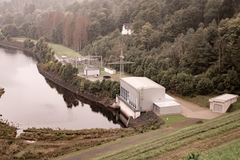 A facility is located near the edge of a large body of water, surrounded by dense, lush green forest trees on all sides. The facility appears to be an industrial or utility building with adjoining smaller structures, power lines, and electrical equipment. There is a road leading into the facility. Behind the forest, a hilly region extends into the background, and a single white house can be seen atop a hill. The water body has a calm and reflective surface.