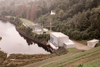 A facility is located near the edge of a large body of water, surrounded by dense, lush green forest trees on all sides. The facility appears to be an industrial or utility building with adjoining smaller structures, power lines, and electrical equipment. There is a road leading into the facility. Behind the forest, a hilly region extends into the background, and a single white house can be seen atop a hill. The water body has a calm and reflective surface.
