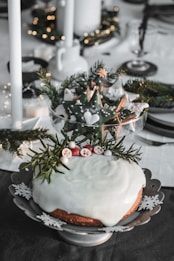 A beautifully decorated holiday cake with white icing topped with sprigs of rosemary and small red and white fruits sits on an ornate plate featuring snowflake patterns. In the background, a bowl of star-shaped cookies decorated with green icing and white patterns is displayed. Evergreen branches and warm white lights add a festive touch to the setting, which includes candles and elegant tableware.
