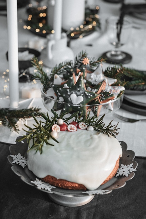 A beautifully decorated holiday cake with white icing topped with sprigs of rosemary and small red and white fruits sits on an ornate plate featuring snowflake patterns. In the background, a bowl of star-shaped cookies decorated with green icing and white patterns is displayed. Evergreen branches and warm white lights add a festive touch to the setting, which includes candles and elegant tableware.
