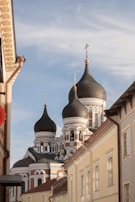 The image captures an architectural scene with prominent onion domes typical of Russian Orthodox churches, adorned with golden crosses. The building is seen from below, contrasting against a clear blue sky with wispy clouds. Flanking the church are residential buildings with pastel-colored facades.