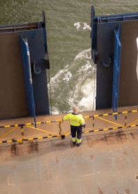 A person in a high-visibility jacket stands on the deck of a vessel, observing the water as seagulls fly over the waves. The deck is bordered by a rail with yellow and black markings, and large metal structures frame the view of the sea.