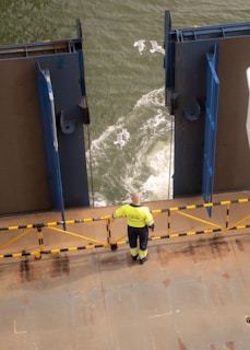 A person in a high-visibility jacket stands on the deck of a vessel, observing the water as seagulls fly over the waves. The deck is bordered by a rail with yellow and black markings, and large metal structures frame the view of the sea.